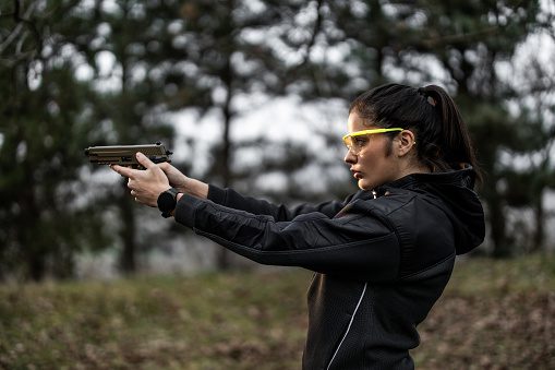 Woman aiming a firearm outdoors, wearing safety glasses and a jacket for secure outdoor training.